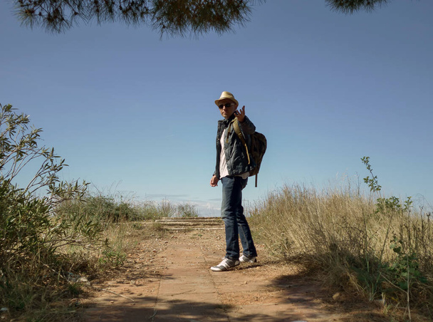 Um homem viajante em um chapéu e com uma mochila fica no convés de observação de uma colina e estende a mão
 - Foto, Imagem