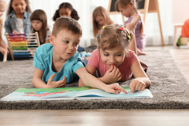 Χαριτωμένα παιδιά διαβάζουν το βιβλίο στο πάτωμα, ενώ άλλα παιδιά παίζουν μαζί στο νηπιαγωγείο - Φωτογραφία, εικόνα