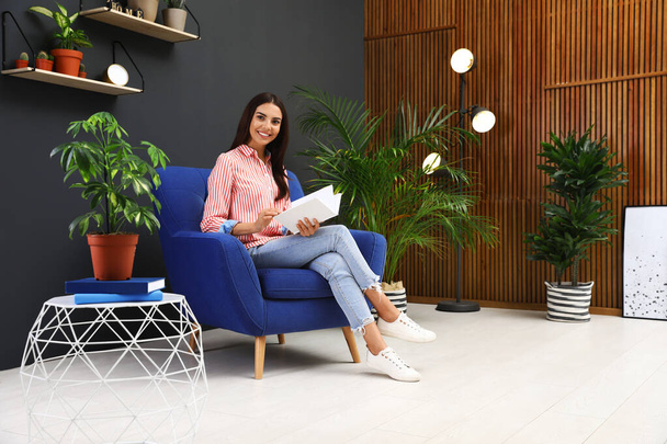 Happy woman reading book in armchair at home. Indoor plants for trendy interior design - Foto, imagen