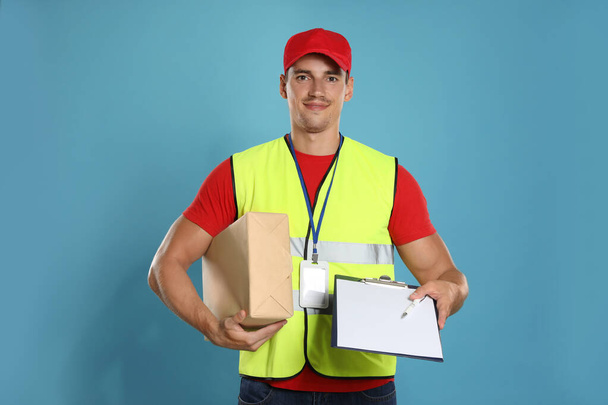 Happy young courier with parcel and clipboard on blue background - 写真・画像