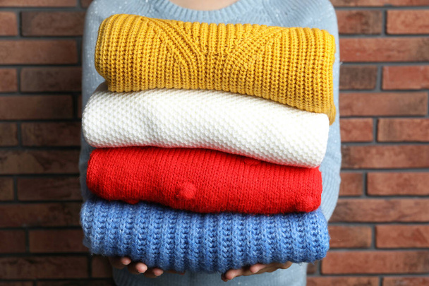 Woman holding pile of winter sweaters near brick wall, closeup view - Фото, изображение