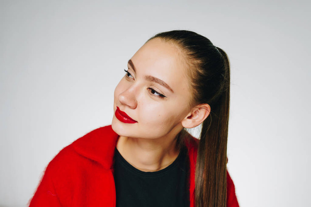 Amazing young girl with dark long hair tied in a ponytail, with red lipstick and in a red sweater with a black T-shirt on a white background, examines something aside with interest - Foto, Imagem