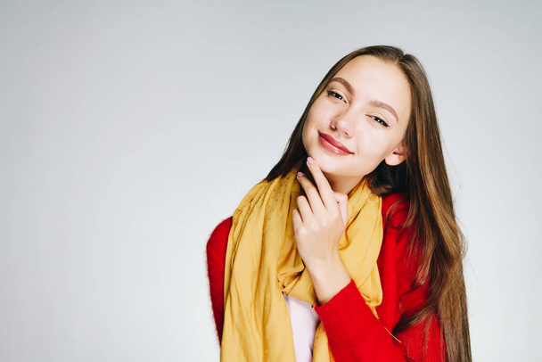 Attractive young girl with dark long hair in a red sweater with a yellow scarf on a white background is up to something - Photo, image