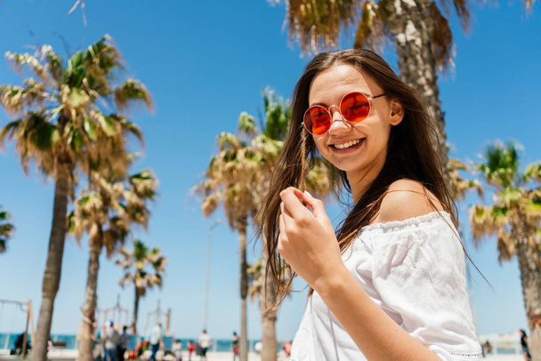 Lovely young girl with long dark hair in the white T-shirt and orange sunglasses smiles against the background of palm trees on a sunny day - Фото, изображение