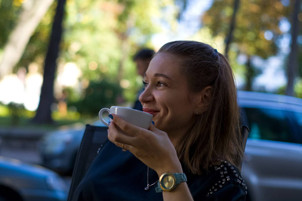 La joven se sienta en un café de verano y bebe café con taza en la mano.. - Foto, Imagen