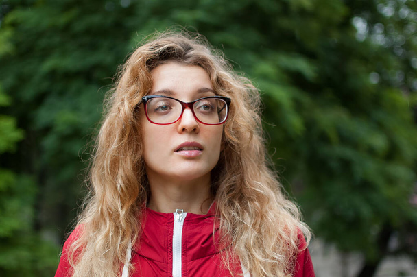 Young beautiful woman with blonde curly hair in dark red hipster jacket and eyeglasses posing outdoors in the urban park in the morning - Photo, Image