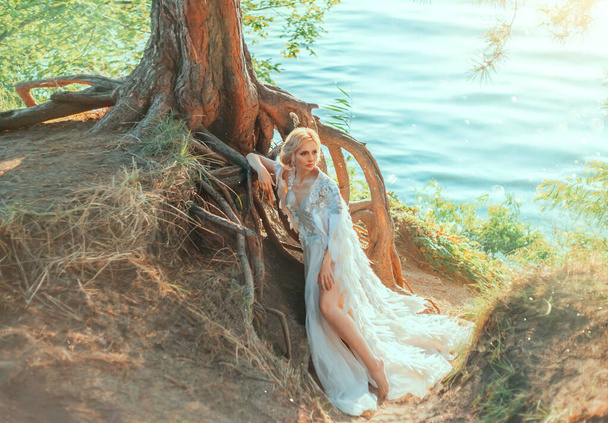 Lujosa mujer rubia en un vestido blanco con una larga capa real con plumas. Queen Swan se encuentra cerca de un árbol con enormes raíces en la costa del río. Fondo verano naturaleza paisaje, colores verdes cálidos
. - Foto, Imagen
