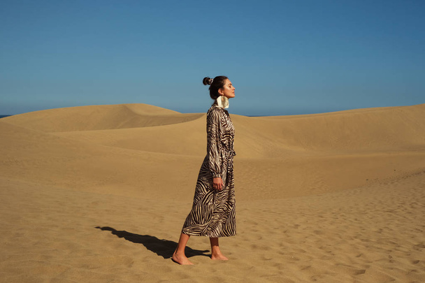 Μοντέλο φορώντας φόρεμα animal print περπάτημα στην αμμώδη έρημο. - Φωτογραφία, εικόνα