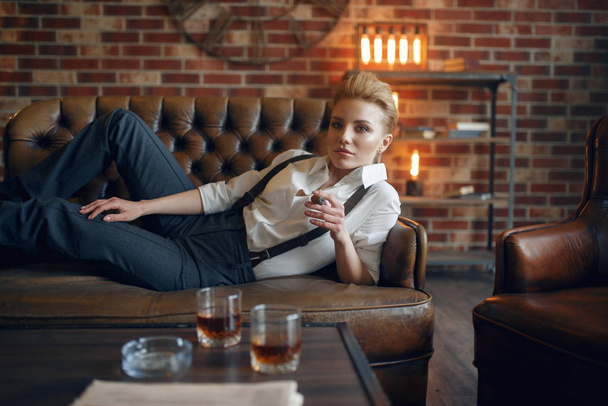 Woman in strict clothes lying on leather couch and smoking cigar, retro fashion, gangster style. Vintage business lady in office with brick walls - Photo, Image