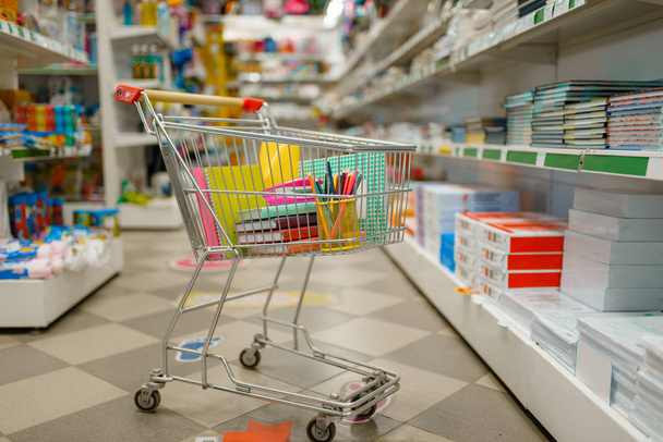Cart with office supplies between the shelves in stationery store, nobody. Assortment in shop, rows of accessories for drawing and writing, school equipment - Fotoğraf, Görsel