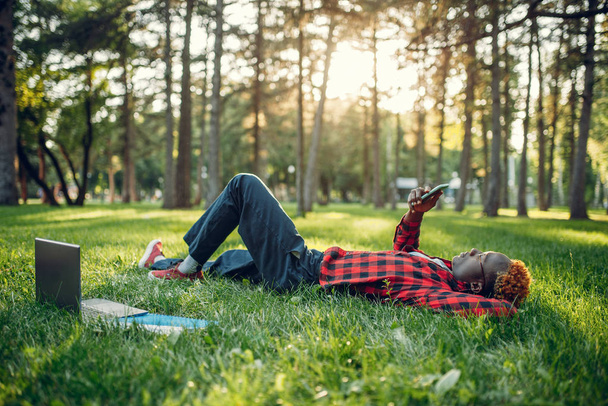 Αφρικανός φοιτητής με τηλέφωνο ξαπλωμένος στο γρασίδι στο καλοκαιρινό πάρκο. - Φωτογραφία, εικόνα