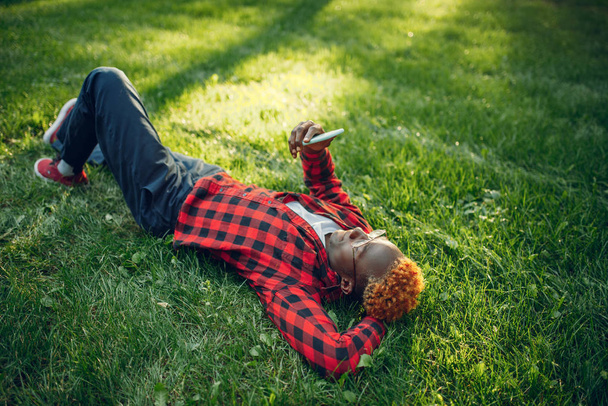 Black student with phone lying on the grass in summer park. A teenager studying and leisures outdoors - Photo, image