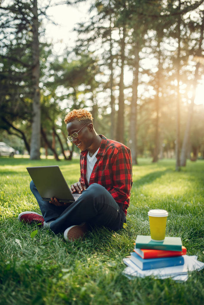 Estudante preto com laptop sentado na grama no parque de verão. Um adolescente estudando e lazeres ao ar livre
 - Foto, Imagem