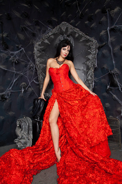 hermosa mujer vampiro en vestido largo rojo cerca del gran trono negro en el estudio
 - Foto, Imagen