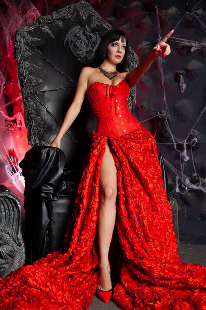 piękna wampirzyca w czerwonej długiej sukience w pobliżu wielkiego czarnego tronu w studio - Zdjęcie, obraz