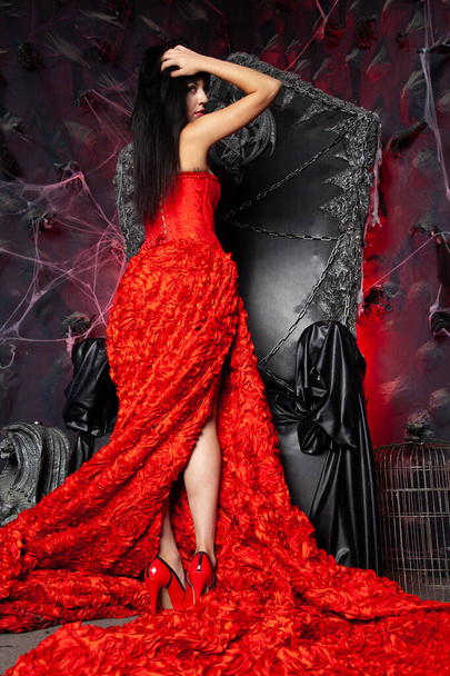 piękna wampirzyca w czerwonej długiej sukience w pobliżu wielkiego czarnego tronu w studio - Zdjęcie, obraz
