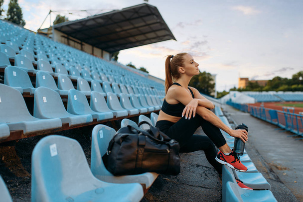 Corredor femenina en ropa deportiva sentada en tribuna y bebe agua, entrenando en el estadio. Mujer haciendo ejercicio de estiramiento antes de correr en arena al aire libre
 - Foto, imagen