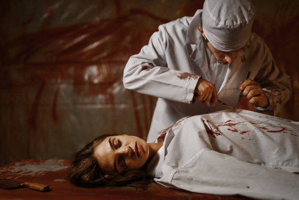 Crazy male psychiatrist with axe and female patient in mental hospital basement, bloody walls on background. Victim of a maniac lying on surgical table in clinic for the mentally ill - Photo, Image