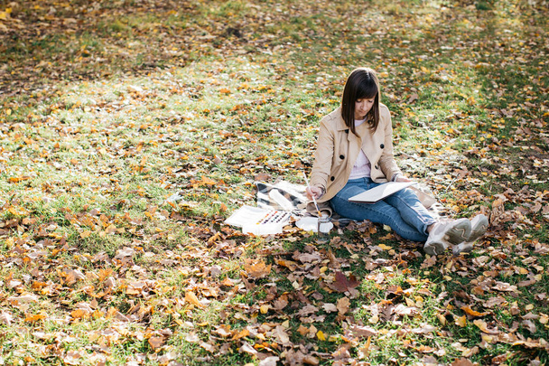 Ένα νεαρό κορίτσι ζωγραφίζει κοντά σε μια λίμνη το φθινόπωρο στο δάσος. Σκιτσάρω. Το κορίτσι έχει καλή διάθεση και είναι χαρούμενο. - Φωτογραφία, εικόνα