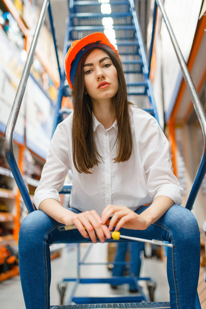 Female customer in helmet sitting on the stairs in hardware store. Buyer look at the goods in diy shop, shopping in building supermarket - 写真・画像