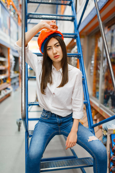 Female customer in helmet sitting on the stairs in hardware store. Buyer look at the goods in diy shop, shopping in building supermarket - Fotó, kép