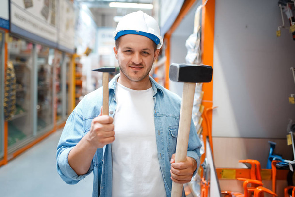 Hombre comprador en casco blanco sosteniendo dos martillos en ferretería. El cliente mira las mercancías en la tienda de bricolaje, compras en el supermercado del edificio
 - Foto, imagen