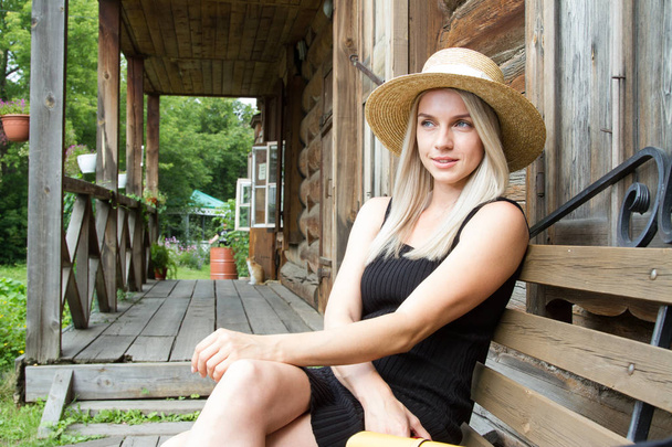 Young blonde in a black short dress sits on a bench by the old log house - 写真・画像
