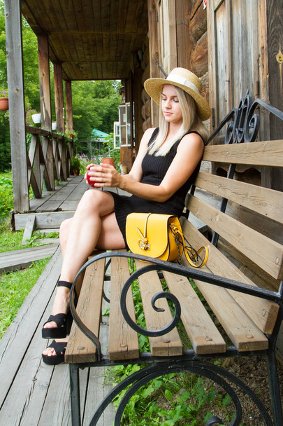 Siyah kısa elbiseli genç sarışın eski ahşap evin yanındaki bankta oturuyor. - Fotoğraf, Görsel