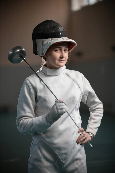A young woman fencer with an open helmet on standing in the gym - Foto, afbeelding