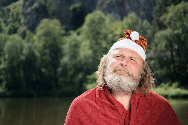 Porträt eines älteren und charismatischen Mannes mit Bart, Weihnachtsmann-Hut und rotem Mantel, der nach oben schaut. das Konzept eines Feiertags oder Urlaubs für Weihnachten und Neujahr in warmen Ländern. Kopierraum. - Foto, Bild