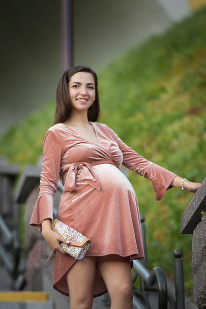 Zwanger meisje poseert op de trap. brunette in een fluwelen jurk. in de open lucht - Foto, afbeelding