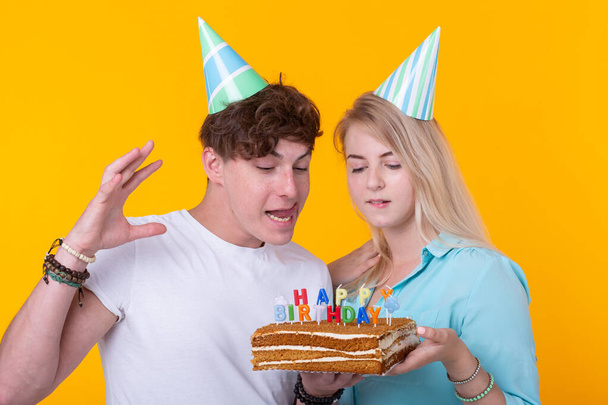 Srandovní muž a žena nosí svátky a brýle držící narozeninový dort se svíčkami na žlutém pozadí - Fotografie, Obrázek