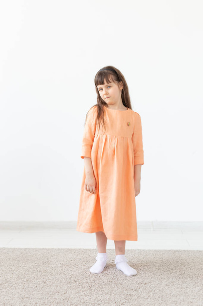 Красивая маленькая девочка в персиковом платье позирует против белой стены. Концепция детской дизайнерской одежды
. - Фото, изображение