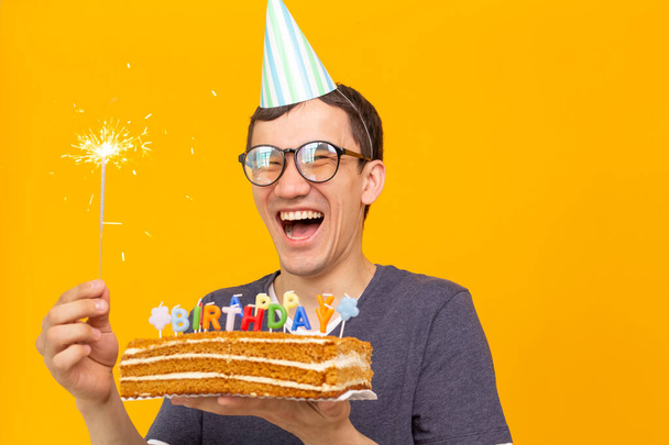 Crazy cheerful young man in glasses and paper congratulatory hats holding cakes happy birthday standing on a yellow background. Jubilee congratulations concept. - Photo, Image