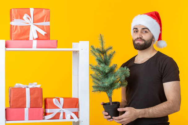 Jonge positieve man met een baard en kerstman hoed staat in de buurt van stand met geschenken en kerstboom op gele achtergrond. Concept van felicatie geschenk en kortingen voor Kerstmis en Nieuwjaar. - Foto, afbeelding