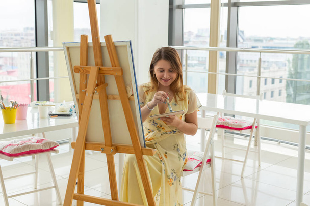 Art school, creativity and leisure concept - student girl or young woman artist with easel, palette and paint brush painting picture at studio - Photo, Image