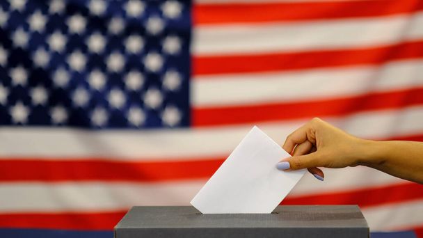 женщина положила бюллетень в урну для голосования в день выборов. Закройте руки белой бумагой для голосования на фоне американского флага
. - Фото, изображение
