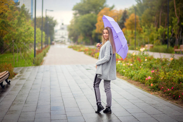 Uma mulher de calças cinza com um guarda-chuva lilás caminha no parque de outono
 - Foto, Imagem