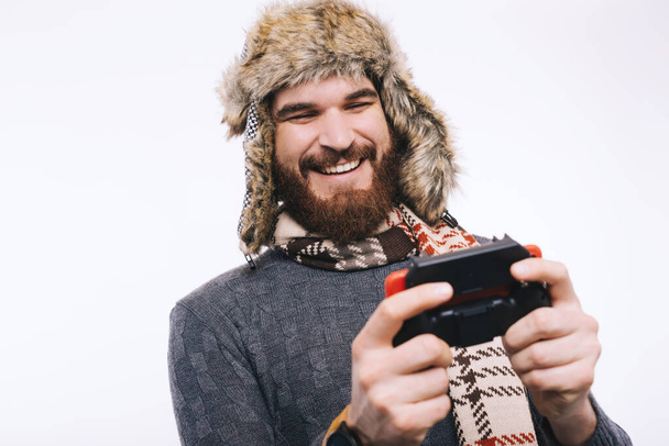 Happy bearded man wearing a winter fur cap hat, a scarf and a sweater playing with a joystick, smiling and being happy. - Photo, Image