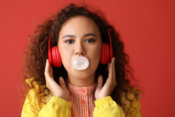 Beautiful African-American woman with chewing gum listening to music on color background - Photo, Image