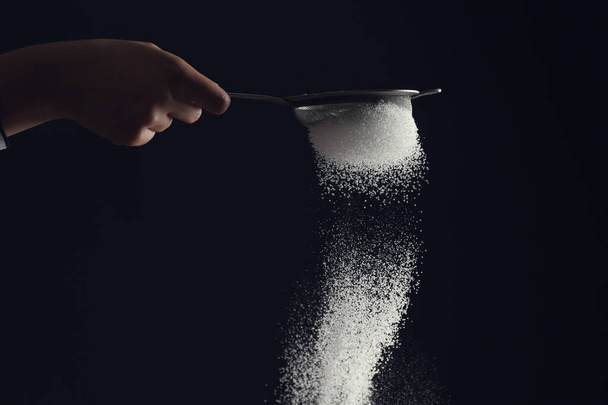 Nainen ripottelee sokeria jauhetta seulan läpi tummalla pohjalla
 - Valokuva, kuva