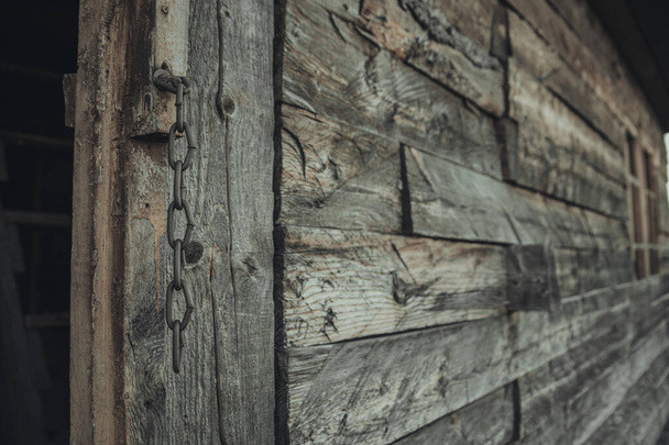 Wooden wall of an abandoned old house. In place of the door is fixed a thick chain. The chain in focus in the foreground the wall fades into the background in a blur. - Fotografie, Obrázek