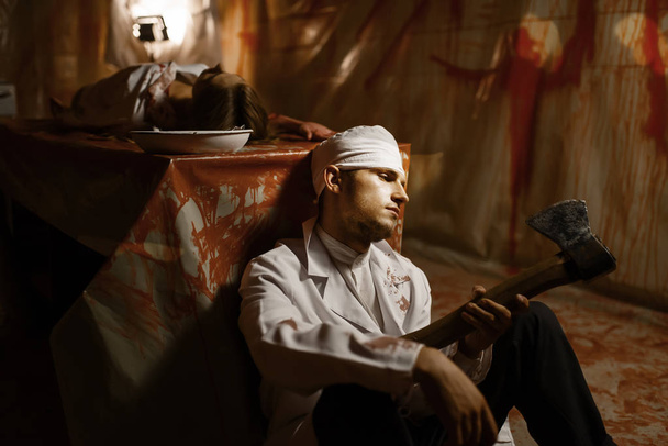 Szalony psychiatra z tasakiem mięsnym siedzący obok pacjentki, eksperyment w piwnicy szpitala psychiatrycznego, krwawe ściany w tle. Ofiara szalonego lekarza w klinice dla chorych psychicznie - Zdjęcie, obraz