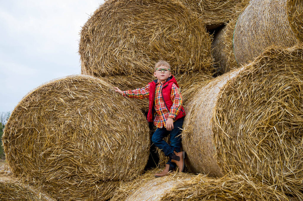 Onnellinen koulupoika heinäsuovassa. Vaalea lapsi punaisessa takissa. Loma ekologisella maatilalla. Maatalouden koulutus. - Valokuva, kuva