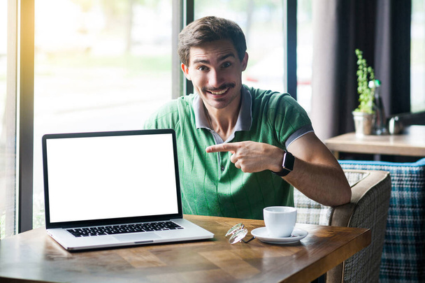 Young businessman in green t-shirt pointing at laptop display and looking at camera with happy face while sitting in cafe, business and freelancing concept - Foto, imagen