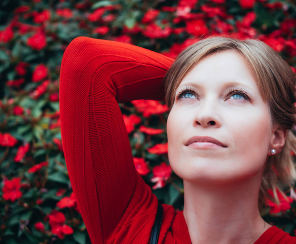 ξανθό κορίτσι με μπλε μάτια και ντυμένο στα κόκκινα, κοιτάζει ψηλά στοχαστικό με φόντο τα κόκκινα λουλούδια - Φωτογραφία, εικόνα
