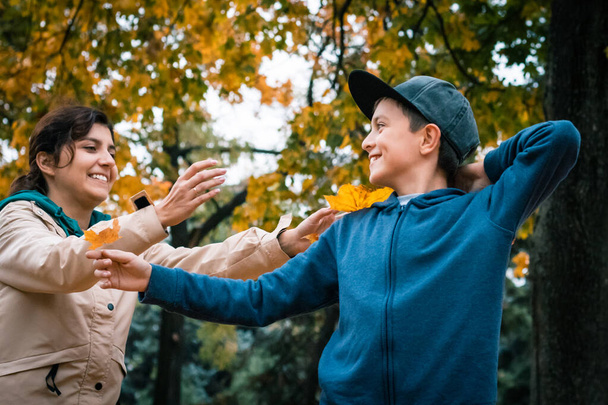 青いパーカーと黒いスナップバックを着た少年がジャンプし、飛んでいる黄色、オレンジ、赤の葉の間で秋の公園で実行されます。カラフルな森と季節の公園のコンセプト. - 写真・画像