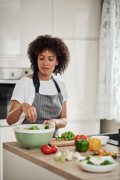 Attractive mixed race woman in apron standing in kitchen and putting chopped lettuce in bowl. Meal preparation concept. - 写真・画像