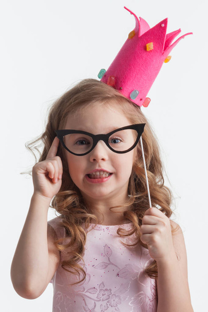 Funny princess girl in pink dress and crown holding party glasses on stick and saying something smart with finger up - Foto, Imagen