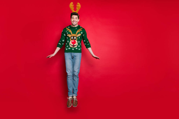 Full body photo of funky guy jumping high excited with x-mas discount prices wear knitted green pullover with ugly deer ornament isolated red color background - Фото, изображение